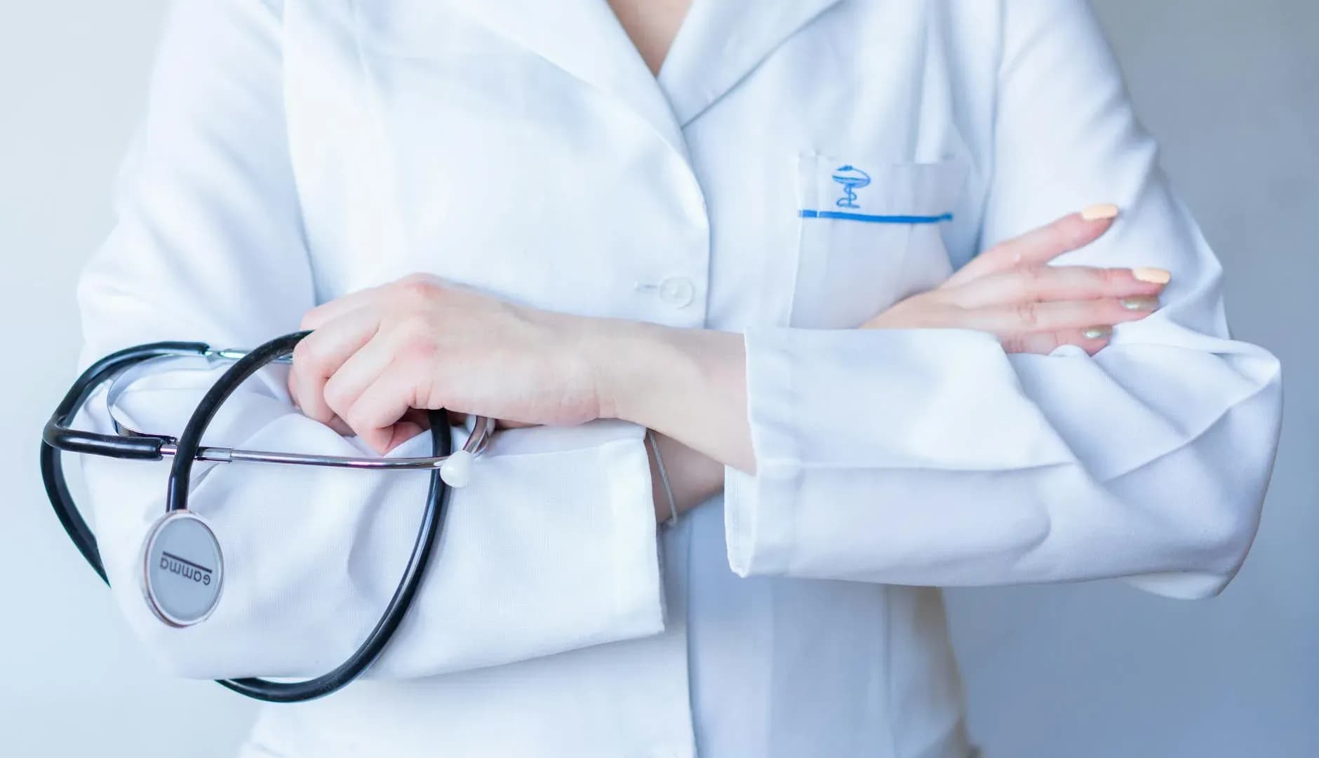 Close-up of a doctor in a white coat with a stethoscope, arms crossed, against a neutral background.